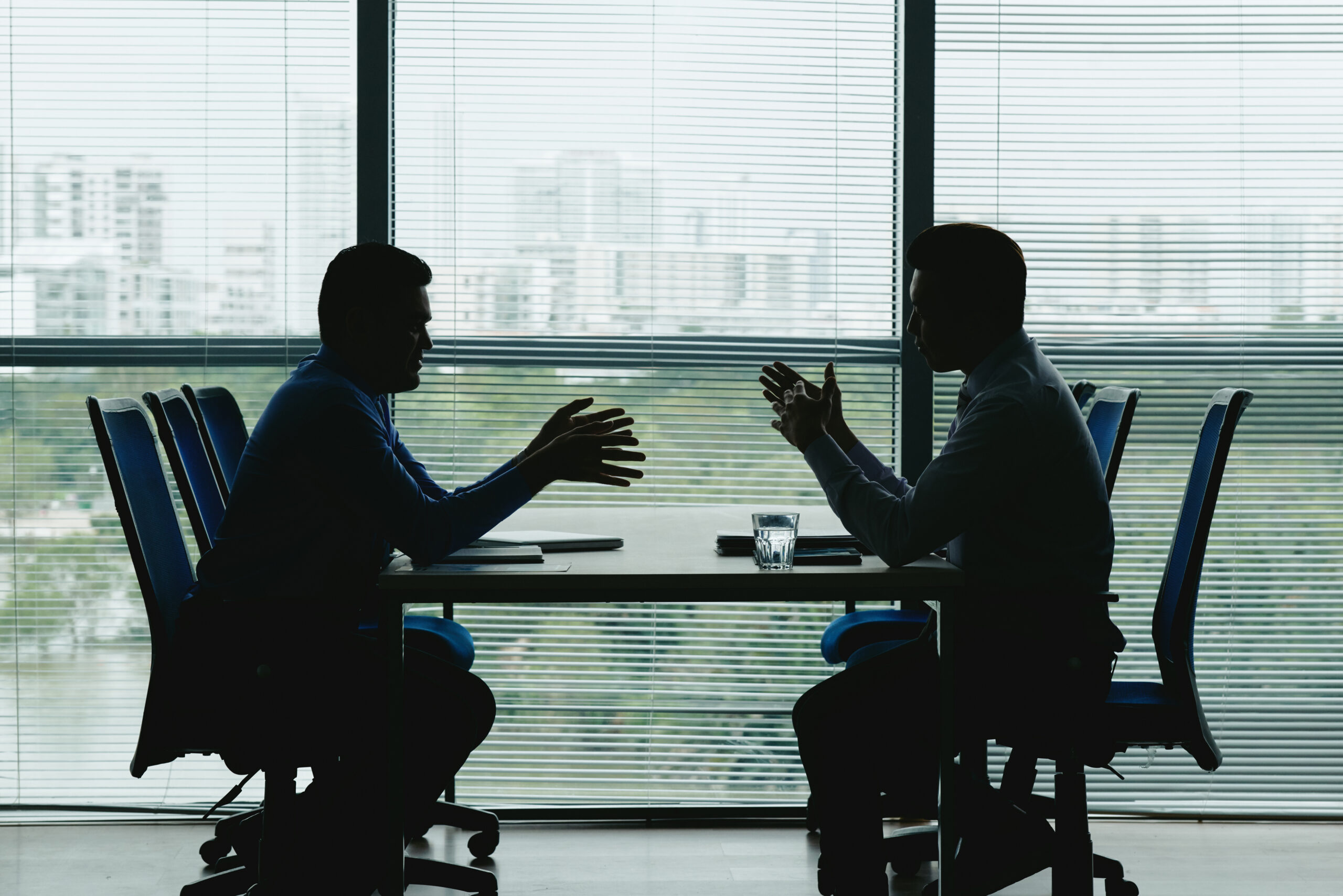 silhouettes-of-two-businessmen-sitting-at-a-table-in-front-of-window-with-view-of-office-park