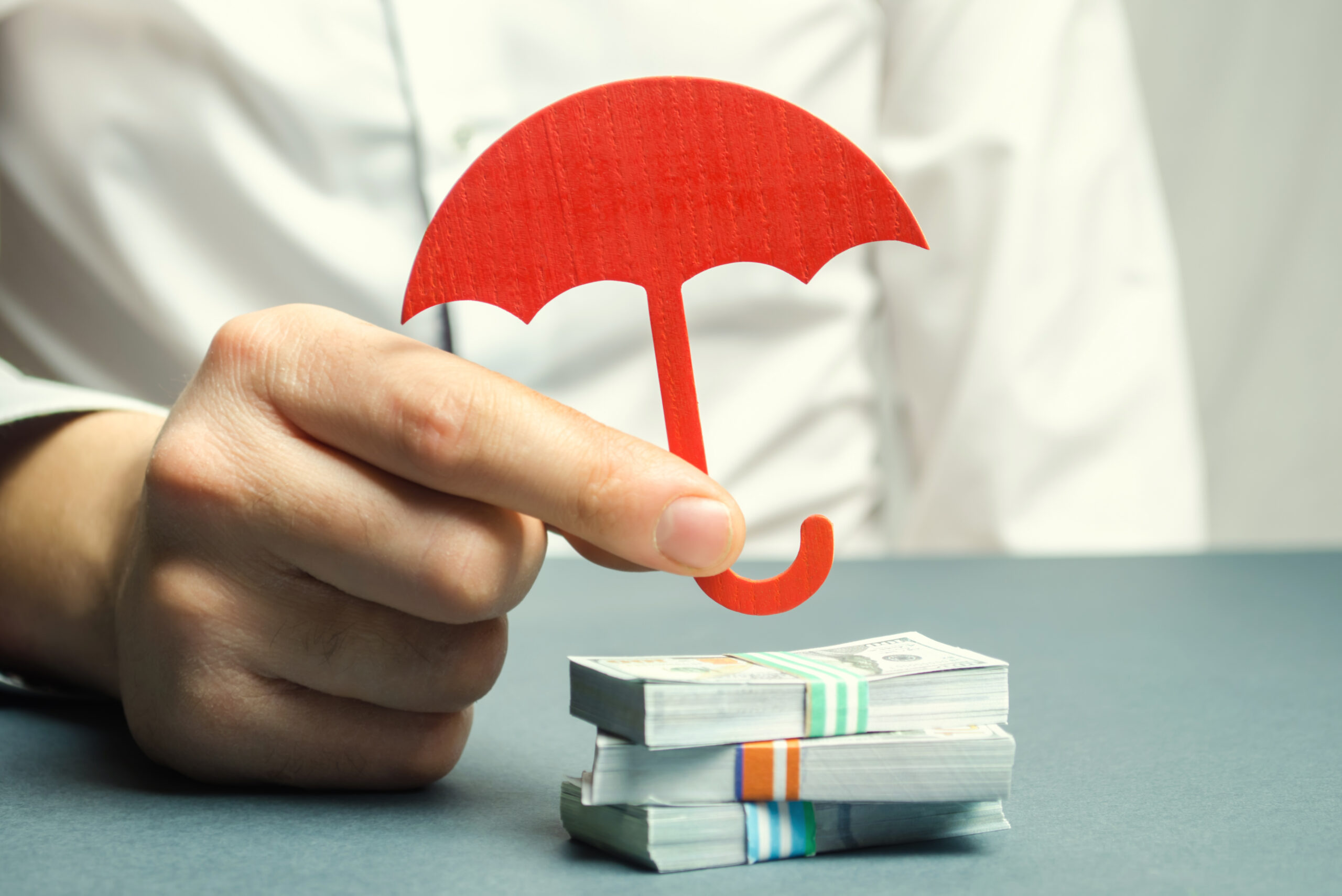 businessman-holding-small-cardboard-cutout-umbrella-over-miniature-bundles-of-money-to-symbolize-protecting-your-personal-assets
